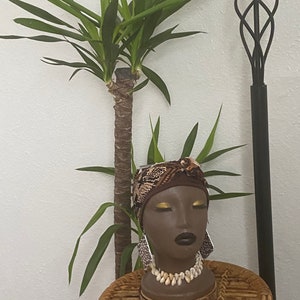 Pode incluir: Uma cabeça de manequim marrom escura com um turbante estampado em marrom e branco, com sombra de olhos dourada e batom escuro. O manequim está usando um colar de conchas brancas e brincos grandes de prata. O manequim está sentado em um banquinho de vime na frente de uma planta verde.