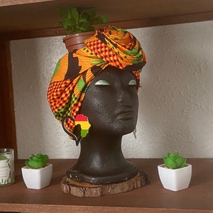 Pode incluir: Um busto de manequim preto é adornado com um turbante vibrante e estampado em laranja, verde e preto. O manequim usa brincos em forma do continente africano. Uma pequena planta em vaso está no topo da cabeça. Velas decorativas e suculentas na prateleira.