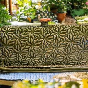 May include: A green ceramic butter dish with a floral pattern. The lid has a small, round handle. The dish is rectangular with a matching base, perfect for serving butter. The design is embossed, adding texture and visual interest.