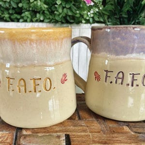 Peut inclure: Deux mugs en céramique avec des anses. L'un est beige avec le haut marron, l'autre est beige avec le haut violet. Les deux mugs portent les lettres "F.A.F.O." et un symbole de main. Les mugs sont sur une surface en bois.