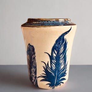 May include: A ceramic vase with a cream-colored base and a flared rim featuring a dark blue glaze. The exterior is decorated with two detailed dark blue feather illustrations printed on the surface, with a smooth, glossy finish throughout.