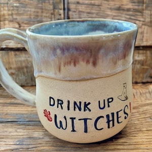 May include: A handmade ceramic mug with a beige base and a blue and maroon rim. The mug features the text "DRINK UP WITCHES!" in dark blue, along with a red hand and a witch's hat illustration. The mug has a large, curved handle.