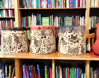 Op de afbeelding: Drie keramische mokken met een crèmekleurige basis en zwarte lijntekeningen van stapels boeken, open boeken, brillen, vlinders en kleine hartjes. Elke mok heeft een afzonderlijk gekleurde rand in donkerbruin, roze of blauw.