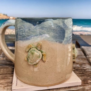 Pode incluir: Uma caneca de cerâmica com um design de esmalte de dois tons, apresentando uma secção inferior em bege areia e uma secção superior azul. Uma tartaruga marinha esculpida em três dimensões está fixada na frente, com outra visível na lateral.