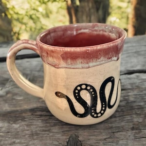 May include: Cream-colored ceramic mug with a black snake design. The rim is a reddish-brown color, and the handle is a matching cream color. The mug is sitting on a wooden surface.