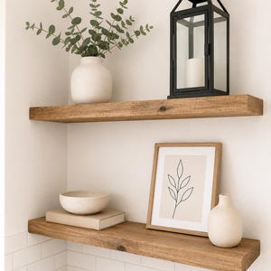 Może przedstawiać: Dwie półki wiszące z naturalnego drewna zamontowane na białej ścianie z płytek. Na górnej półce znajduje się biały wazon z zielonymi gałązkami eukaliptusa oraz czarna metalowa latarnia. Dolna półka zawiera beżową książkę, nakrapianą miskę, oprawiony rysunek liścia oraz mały biały wazon.