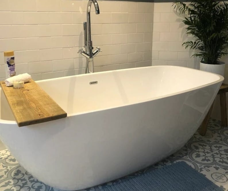 May include: A modern white bathtub with a wooden bath tray holding a rolled white towel and a small candle. A silver faucet and a green plant in a white pot are visible in the background. The bathroom has white tile walls.