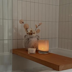 May include: A wooden shelf holds a lit candle in a glass holder, a small candle, and a decorative vase with flowers and foliage. The shelf is set against a backdrop of white tiled walls, creating a spa-like atmosphere.