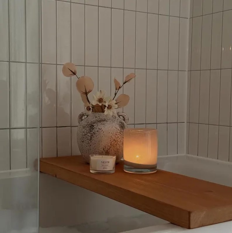 May include: A wooden shelf holds a lit candle in a glass holder, a small candle, and a decorative vase with flowers and foliage. The shelf is set against a backdrop of white tiled walls, creating a spa-like atmosphere.