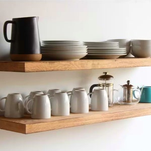 Op de afbeelding: Twee houten planken met keukengerei. De bovenste plank bevat een zwarte kan, borden en kommen. De onderste plank heeft witte mokken, een cafetière, een theepot en een turquoise kan. De items zijn geplaatst tegen een witte muur.