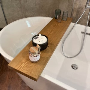 May include: A wooden bathtub tray with a candle, bath salts, and decorative glasses. The tray is made of light-colored wood and spans the width of a white bathtub. A black candle holder and a glass jar with a cork lid are visible.