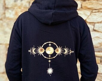 May include: Black hooded sweatshirt with a gold celestial design on the back. The design includes suns, moons, stars, and other celestial elements. The person is wearing blue jeans.