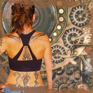 Op de afbeelding: Een vrouw met een rugtattoo met ingewikkelde ontwerpen in zwart en grijs. De tatoeage is zichtbaar door een zwarte sportbeha. De achtergrond is een steampunk-geïnspireerd ontwerp met tandwielen en tandwielen.