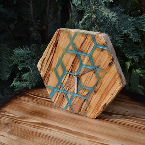 May include: Hexagon shaped wooden clock with a turquoise resin design. The clock has silver hands and is sitting on a wooden surface.