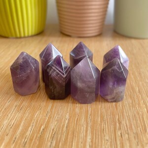 May include: Six small, purple amethyst points arranged in a row on a wooden surface. The points are all roughly the same size and shape, and they have a smooth, polished finish.