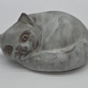 May include: A ceramic figurine of a curled-up cat with a marbled gray and white finish. The cat is depicted in a resting position with its head turned toward the front, featuring dark gray painted eyes, ears, and subtle shading across its body.