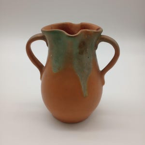 May include: A terracotta pitcher featuring two curved handles and a scalloped rim. The exterior displays a matte orange-brown finish with a decorative green glaze drip pattern extending from the top rim down the sides of the vessel body.