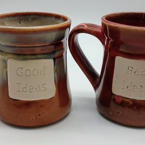 May include: Two ceramic mugs with handles. One is brown with the words "Good Ideas" on a white rectangle. The other is red-brown with "Bad Ideas" on a white rectangle. Both mugs have a glossy finish.