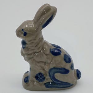 May include: A small ceramic figurine of a rabbit in a seated position. The rabbit has a glossy light brown finish with hand-painted dark blue spots and markings on its ears, back, and sides. The surface is smooth and reflective throughout.