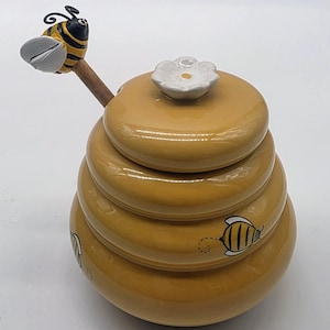 May include: A yellow ceramic honey pot shaped like a beehive with a matching lid topped by a white flower. A wooden honey dipper with a sculpted bee figure on the handle rests inside the pot. Small black and yellow bees are painted on the sides.