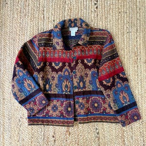 May include: A long-sleeve button-up jacket featuring a multi-colored tapestry pattern with shades of red, blue, gold, and dark brown. The garment has a fold-over collar, a front button closure, and a repeating floral and geometric motif throughout.