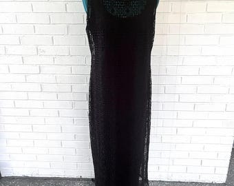 May include: A sleeveless, floor-length black dress. The dress features a delicate lace overlay, with a round neckline and a scalloped hem. The dress is displayed on a mannequin against a white brick wall.