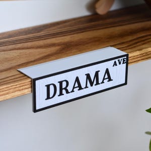 以下が含まれることがあります： 太字の黒い文字で「DRAMA AVE」と書かれた白と黒のサイン。サインは木製の棚に取り付けられています。棚は自然な仕上がりの木材です。装飾品です。