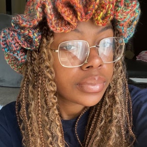 May include: A woman with long, braided hair is wearing a colorful crocheted hat with a ruffled brim. She is also wearing gold-rimmed glasses.