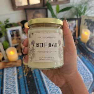 May include: A clear glass jar candle with a gold lid. The label reads "RECUÉRDAME" with scent notes of tangerine, orange, agave, and lime. The candle is 8 oz. and made with all-natural vegan soy wax.