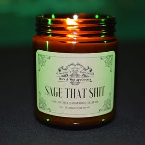 以下が含まれることがあります： 緑色の文字で「SAGE THAT SHIT」と書かれたラベルが付いた、点灯した茶色のガラス瓶キャンドル。ラベルには「Wick & Wax Apothecary」の文字と、セージ、ラベンダー、サンダルウッド、シダーウッドの香りの詳細も記載されています。キャンドルは227gのオールナチュラルビーガンソイワックスです。