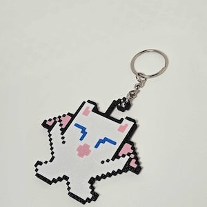 May include: A white pixelated cat keychain with black outlines, pink ears and cheeks, and blue eyes. The keychain is attached to a silver metal ring and chain. The cat is in a standing pose.