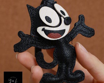 May include: Black leather keychain featuring a cartoon cat with white eyes and a red mouth. The cat is waving with its right paw.