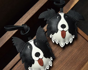 May include: Two black and white leather dog keychains with a stitched border. The keychains are shaped like the head of a border collie with a red tongue.