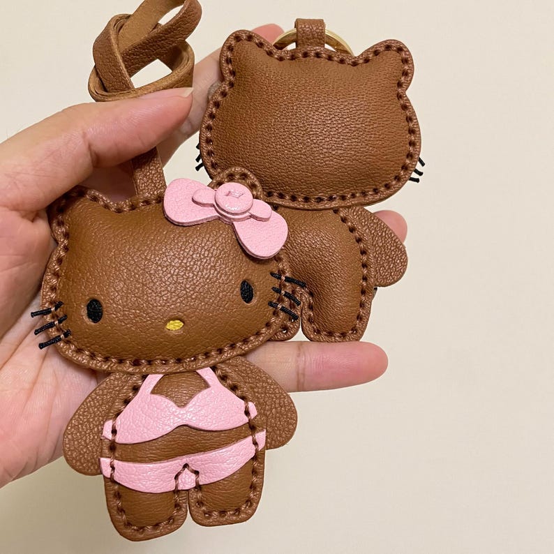 May include: Brown leather Hello Kitty keychain with a pink bikini and bow. The keychain is handmade and features detailed stitching.