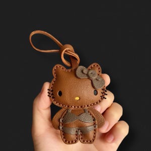 May include: A brown leather Hello Kitty keychain with a brown bow and black stitching. The keychain is in the shape of Hello Kitty and has a brown leather strap.