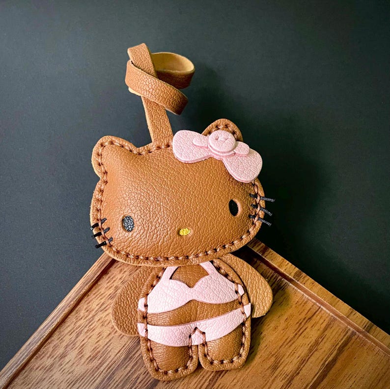 May include: A brown leather Hello Kitty keychain with a pink bow and a pink bikini. The keychain is attached to a brown leather strap.