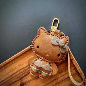 May include: A brown leather Hello Kitty keychain with a gray bow and a gold key ring. The keychain is in the shape of Hello Kitty wearing a gray and brown outfit.