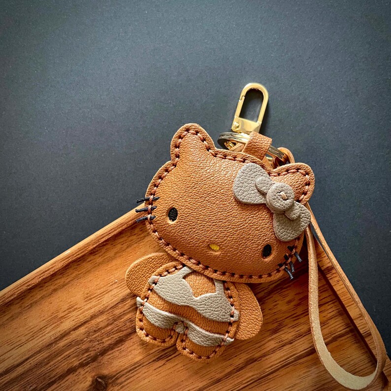 May include: A brown leather Hello Kitty keychain with a gray bow and a gold key ring. The keychain is in the shape of Hello Kitty wearing a gray and brown outfit.
