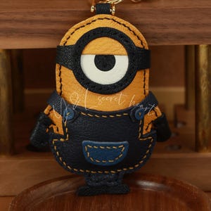 May include: A yellow leather keychain shaped like a minion with a single eye, black goggles, and blue denim overalls. The keychain has a gold metal ring.
