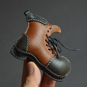 May include: A miniature leather boot in shades of brown and black, with white stitching and black laces. The boot has a rounded toe and a small heel. The leather appears to be textured, and the boot is likely handmade.