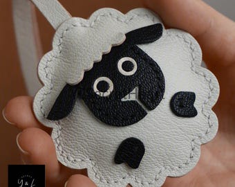 May include: White leather keychain shaped like a sheep with black felt details. The sheep has a black face with two white eyes and a black nose.