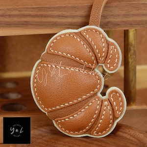 May include: A brown leather croissant keyring with white stitching. The keyring is shaped like a croissant and has a white leather border.