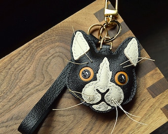May include: Black leather cat keychain with white and orange details. The cat has stitched features and white whiskers.