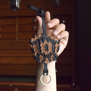 May include: A black and brown leather keychain shaped like a dragon scale. The keychain has a gold ring attached to it.