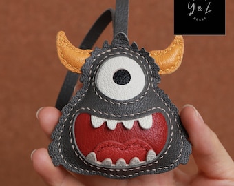 May include: A gray leather monster keychain with a single white eye, red mouth, and yellow horns. The keychain is stitched with white thread and has a black leather strap.
