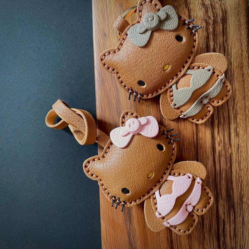May include: Two brown leather keychains shaped like Hello Kitty with pink and gray bows. The keychains have a stitched design and are attached to a brown leather strap.