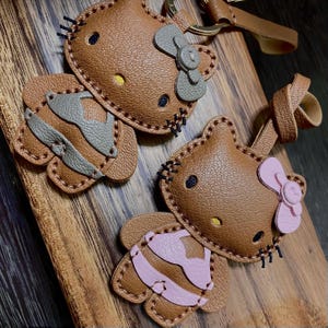 May include: Two brown leather keychains shaped like a cartoon character with a bow. One keychain has a gray swimsuit and the other has a pink swimsuit.
