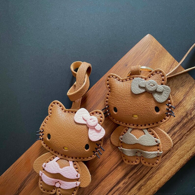 May include: Two brown leather keychains shaped like a cartoon cat wearing a swimsuit. One keychain has a pink swimsuit and a pink bow. The other keychain has a gray swimsuit and a gray bow.
