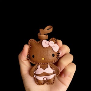 May include: Brown leather Hello Kitty keychain with a pink bow and a pink bikini. The keychain is held in a hand.