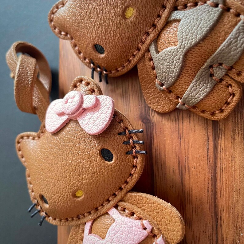 May include: Two brown leather Hello Kitty keychains with pink bows. One keychain has a brown leather bow and the other has a pink leather bow. The keychains are stitched with black thread.